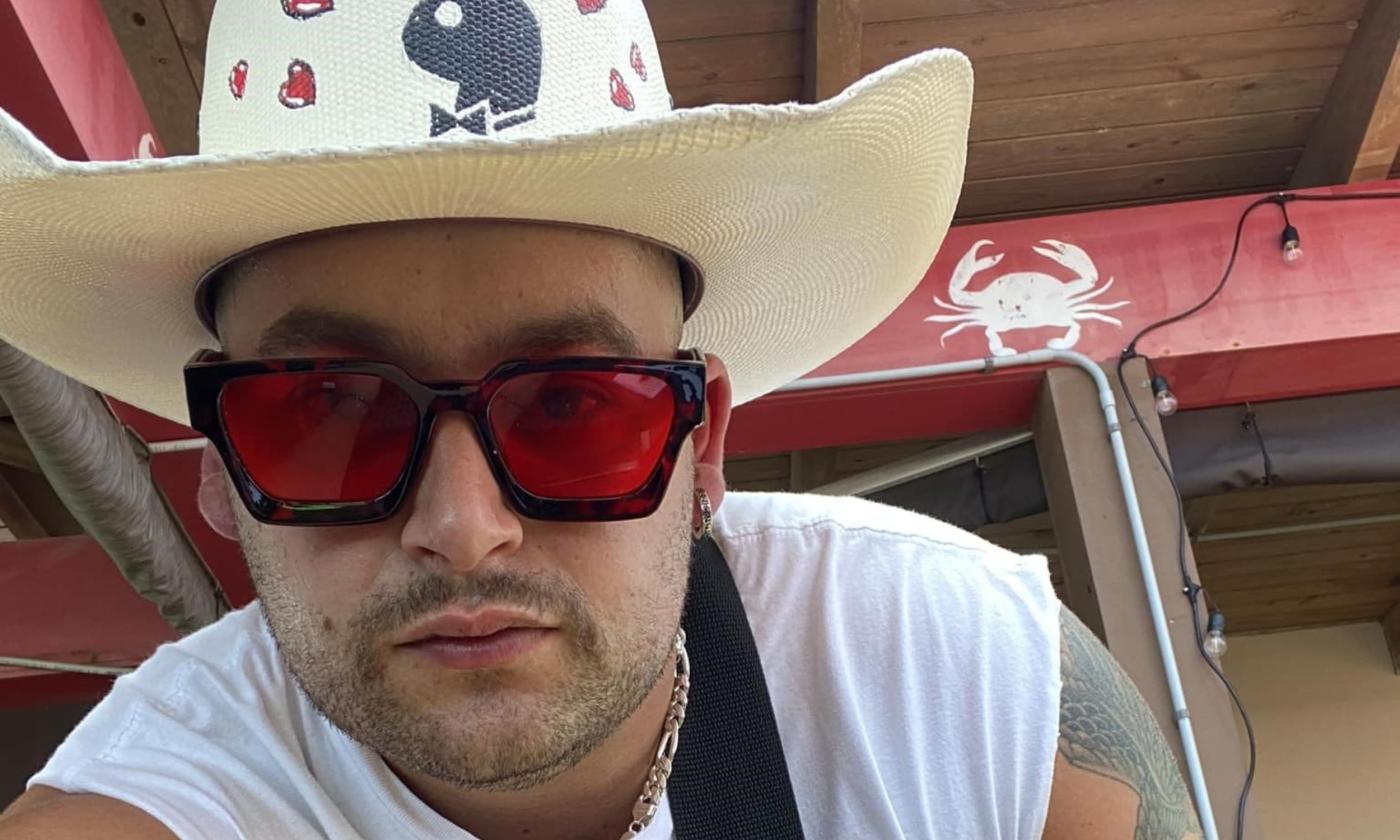 Muddy Marshall poses in a straw hat and red glasses in front of a stage. 