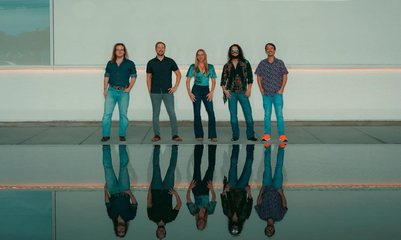 Five musicians pose, standing. Image is split by a body of water on bottom half, on which the musicians are reflected.