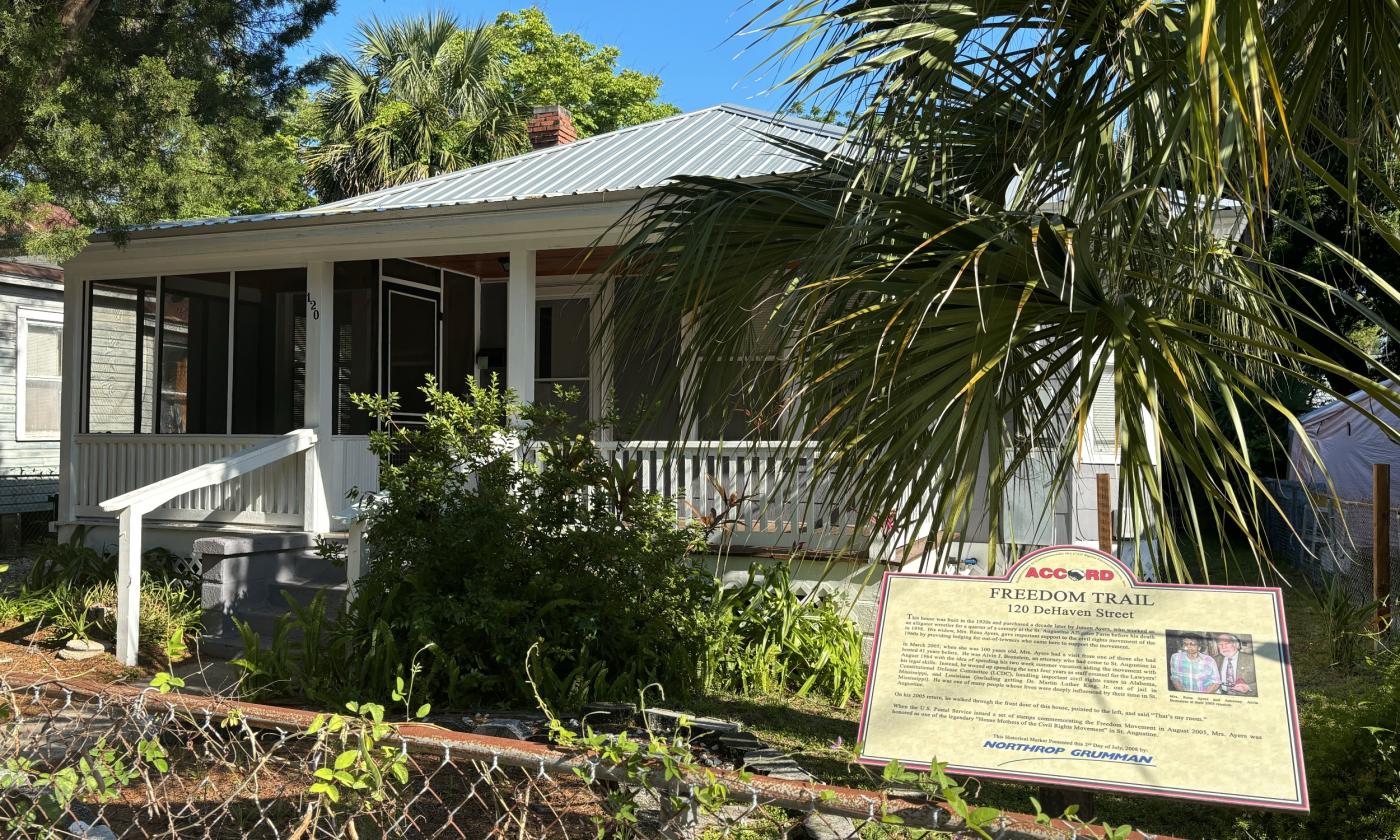 Grey one-story slatboard house with a large front porch, greenery and ACCORD Freedom Trail marker in the foreground.