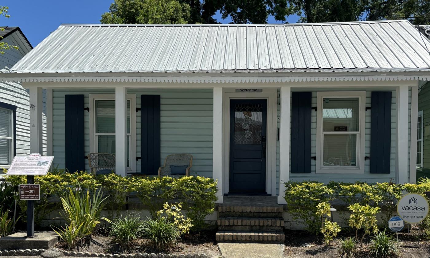 A one-story blue house with a tin roof and white details. An ACCORD Freedom Trail Plaque in front of hedges to the left.