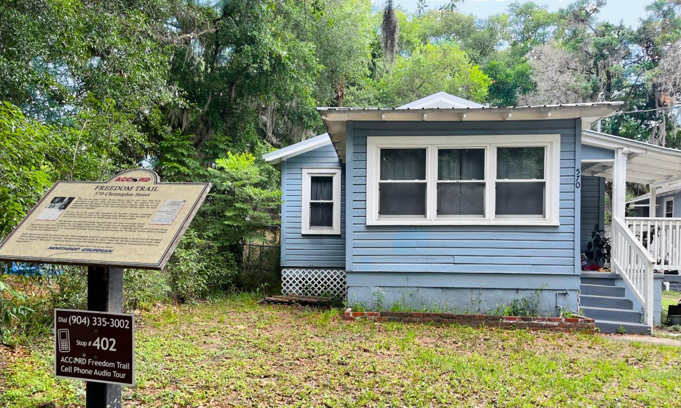 Blue wood slat house backed by greenery, an ACCORD Historical Marker in the foreground.