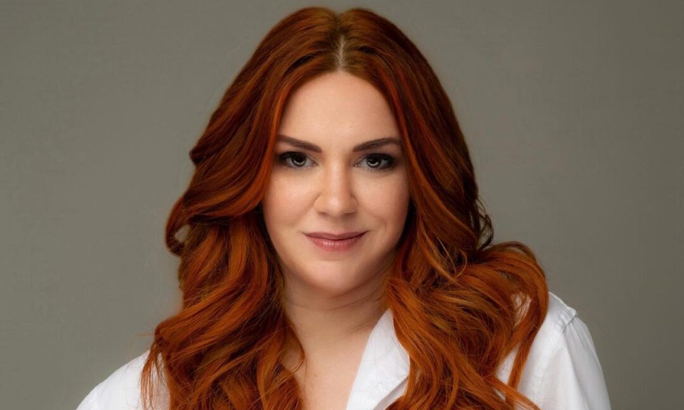 Sarah Ross wears a white blouse while posing in front of a brown backdrop. 