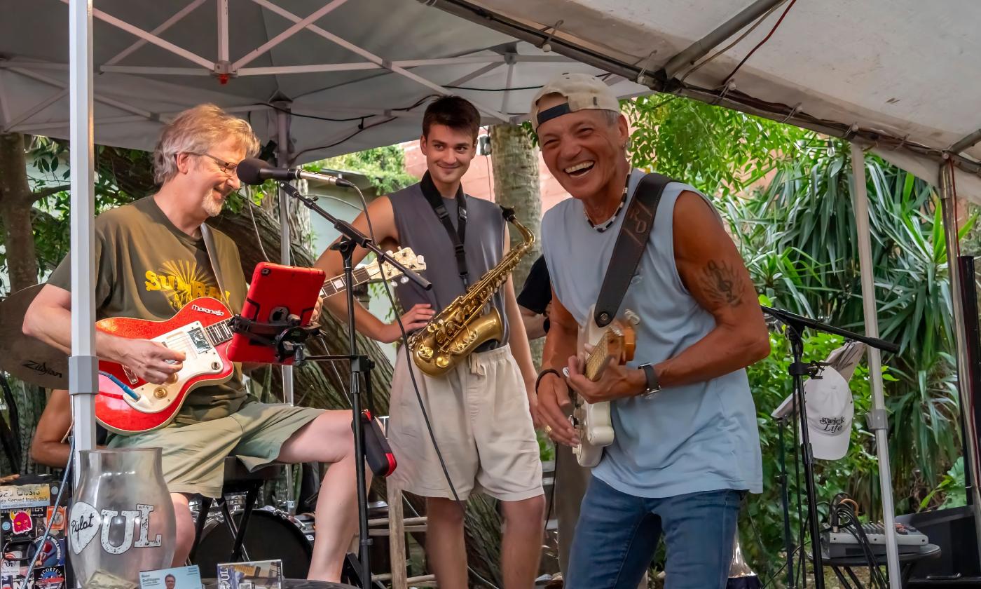 Three musicians, with a drummer in the back, laughing and smiling as they play a song