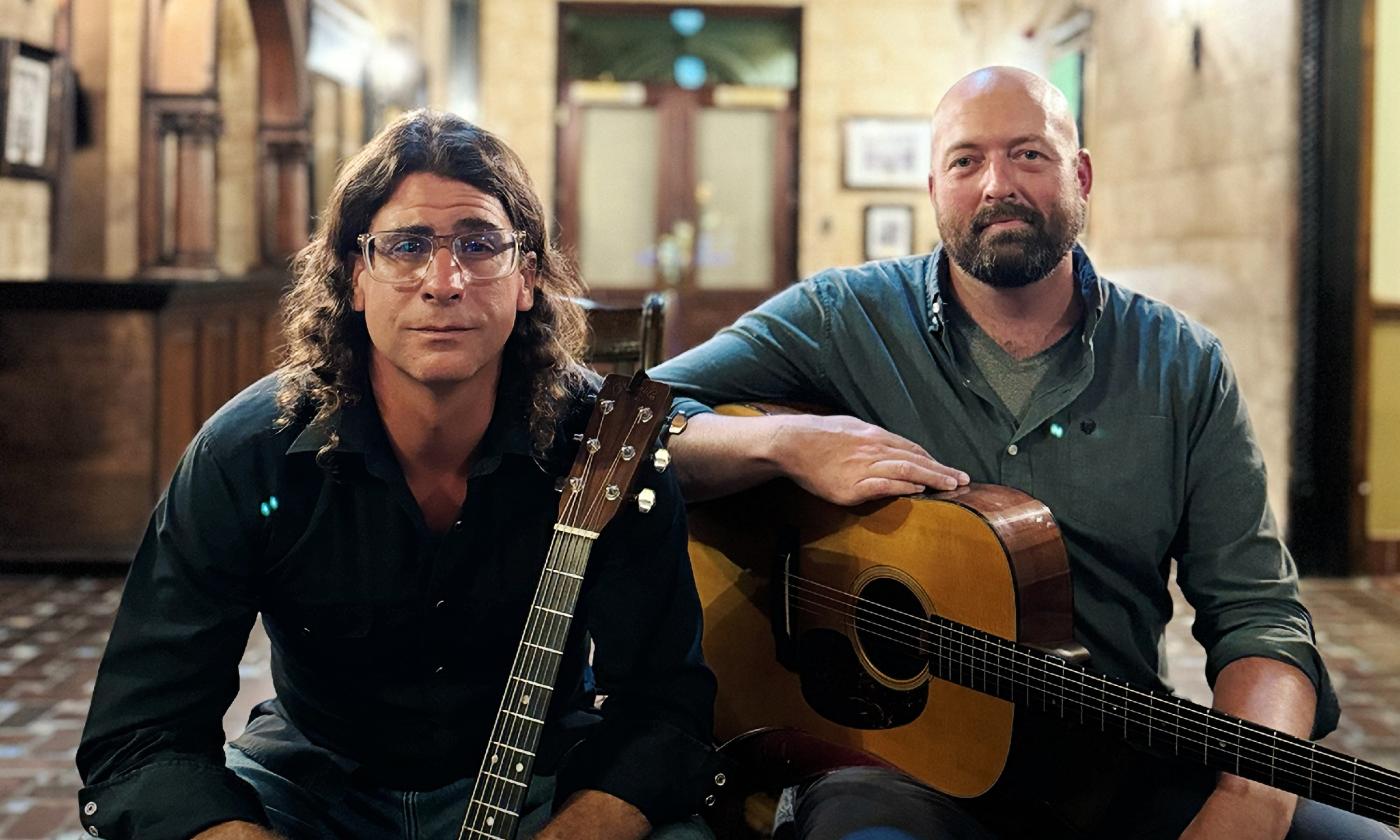 Two men, each holding a guitar, sit in the lobby of an old building in St. Augustine