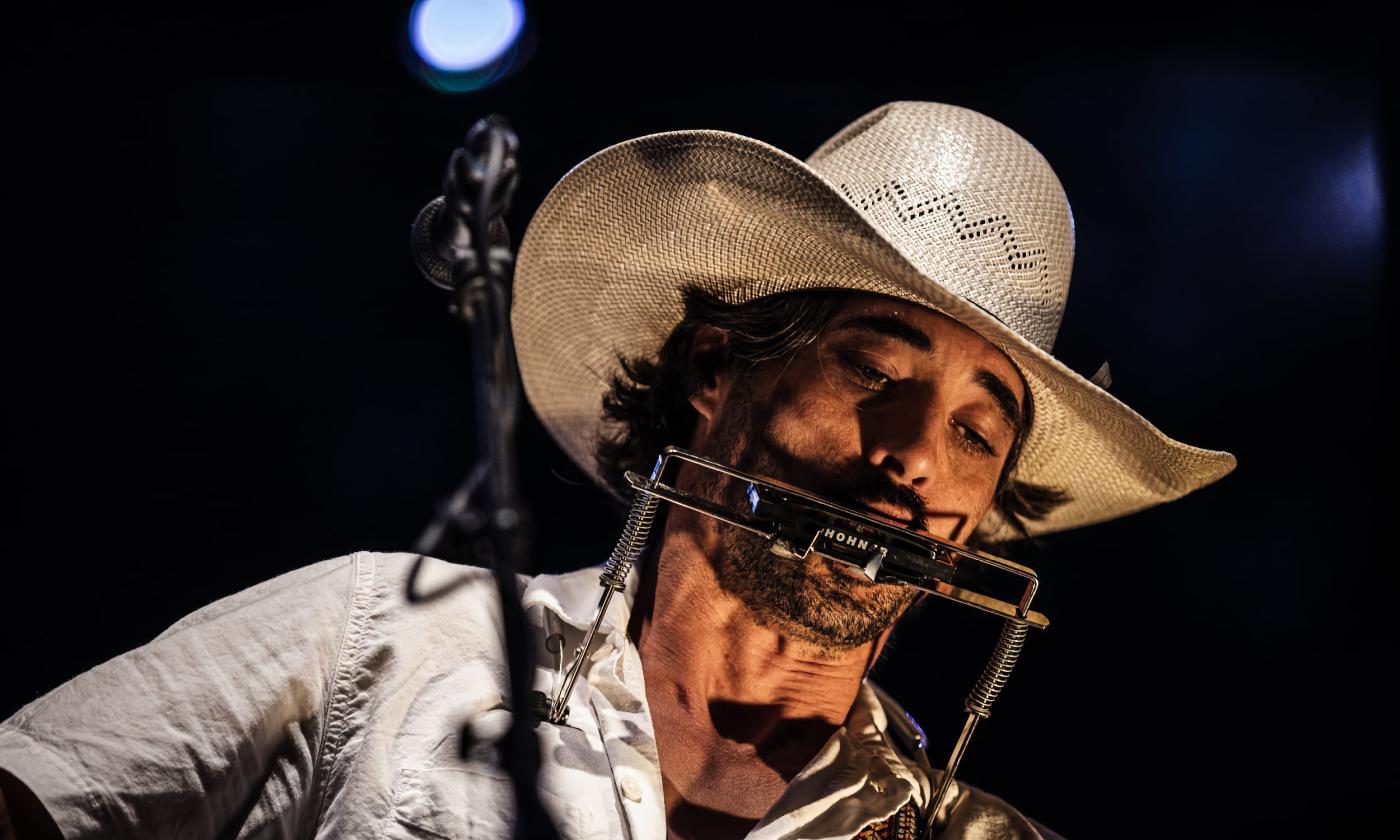 Singer and songwriter Ryan Bingham in white cowboy shirt and hat, on stage with a guitar and harmonica