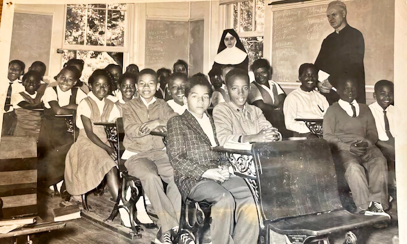 B&W, classroom of young Black students seated at desks, priest and Sister stand behind.