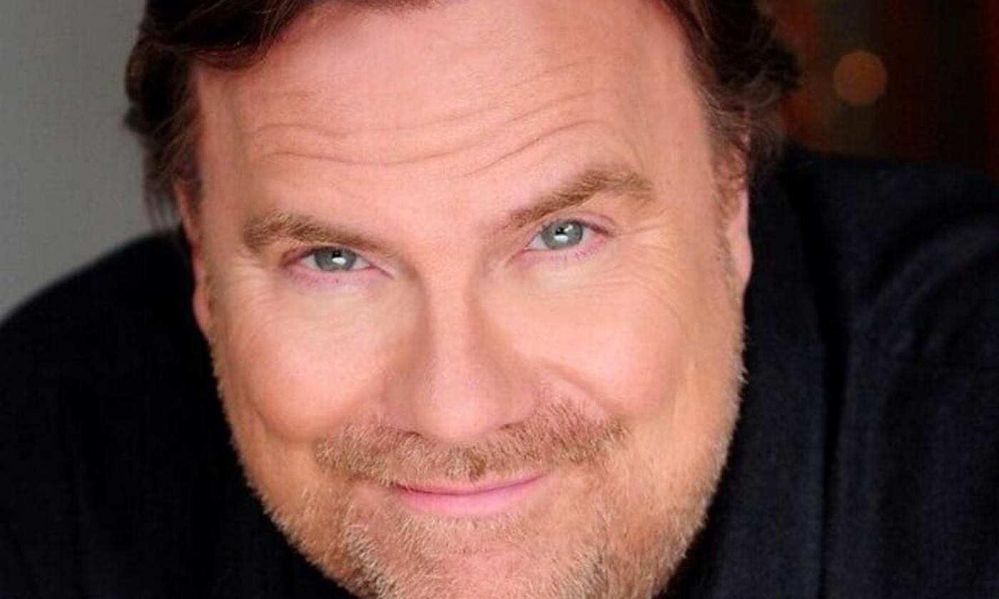 Kevin Farley smiles and poses in front of a grey background with bokeh. 