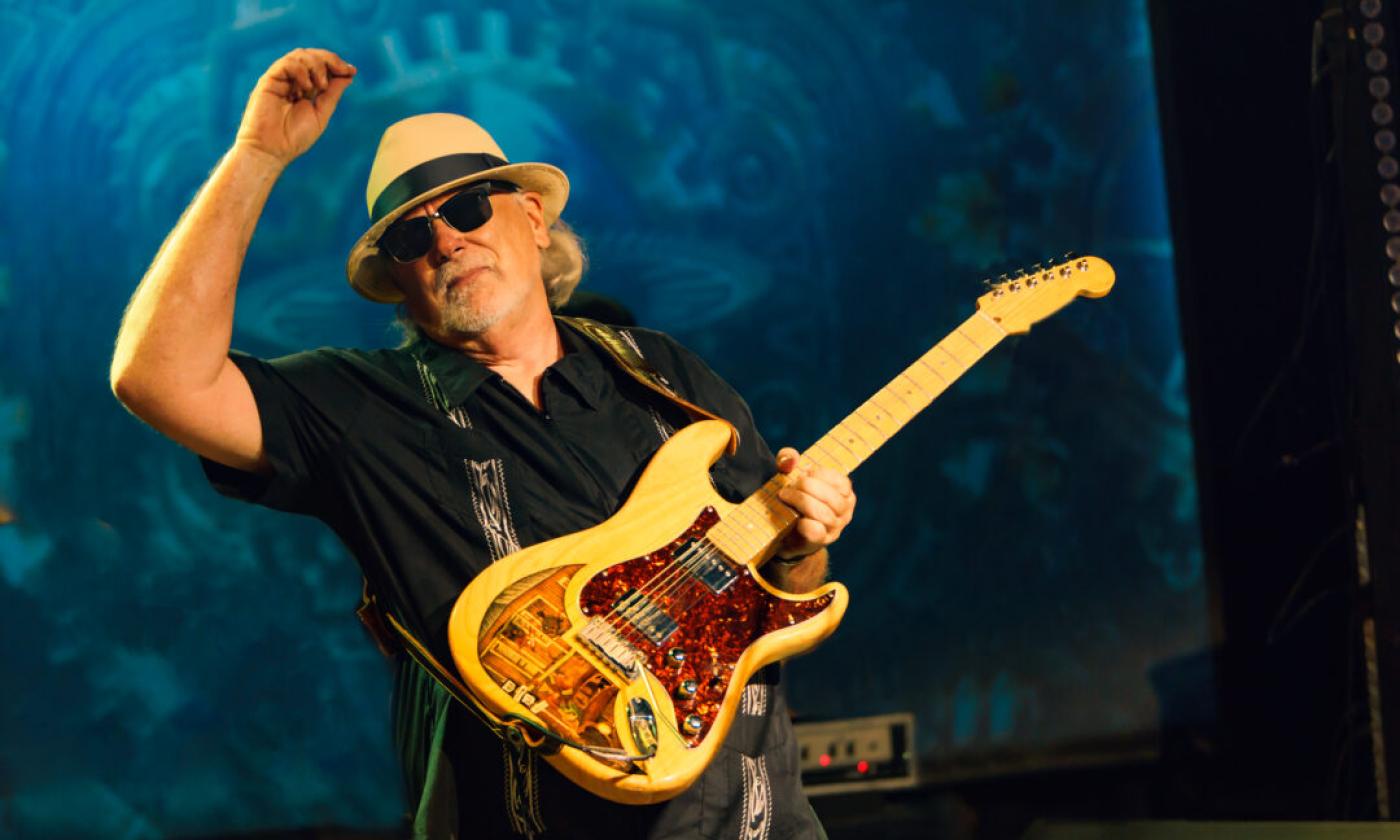 Tas Cru strums his guitar while wearing a hat and glasses. 