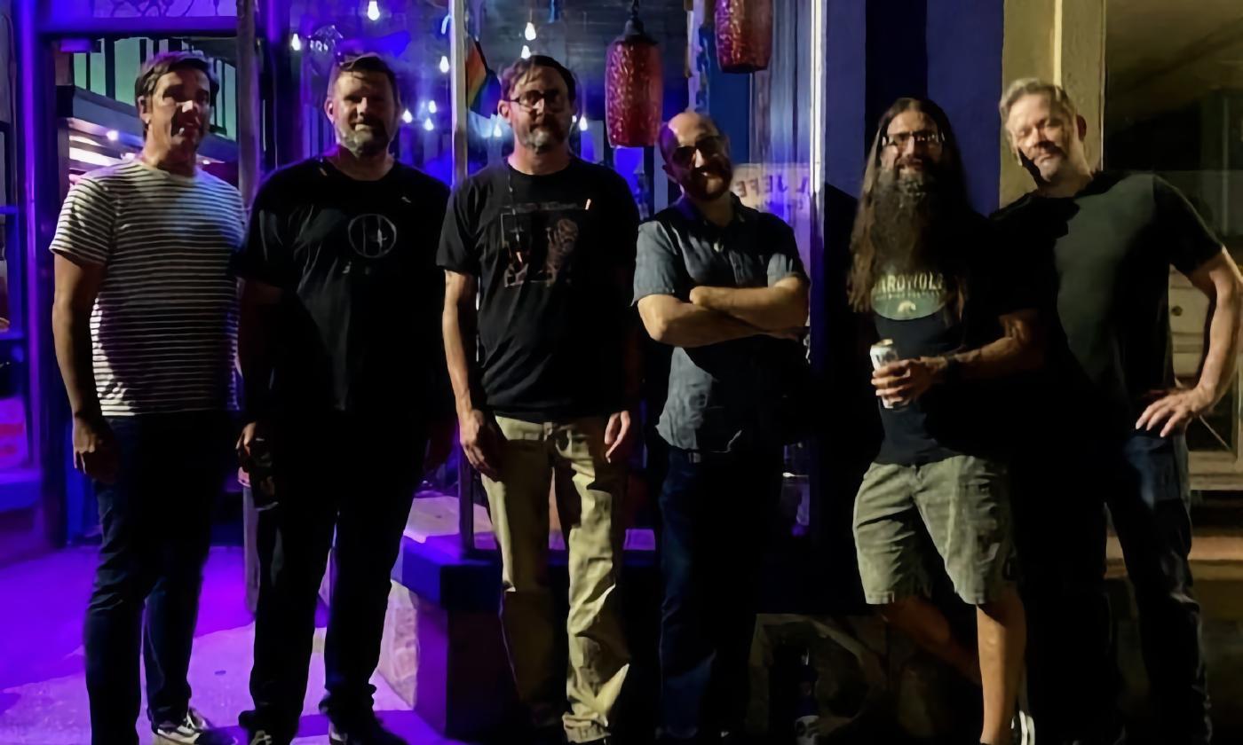 Six men at night, probably members of a band, leanding against a building wall on a corner