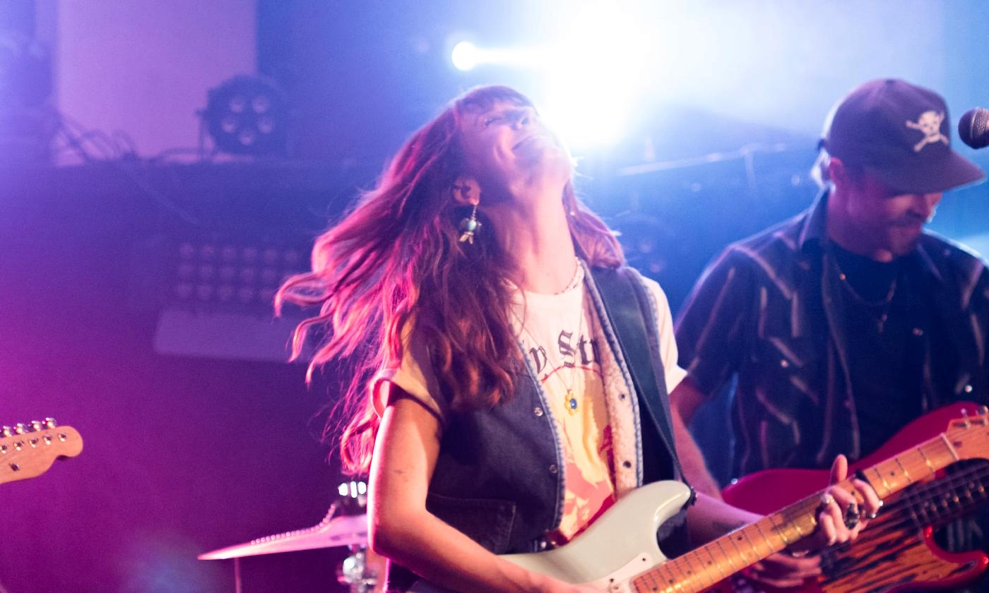 A woman singing on stage, holding a guitar, her hed back in joy