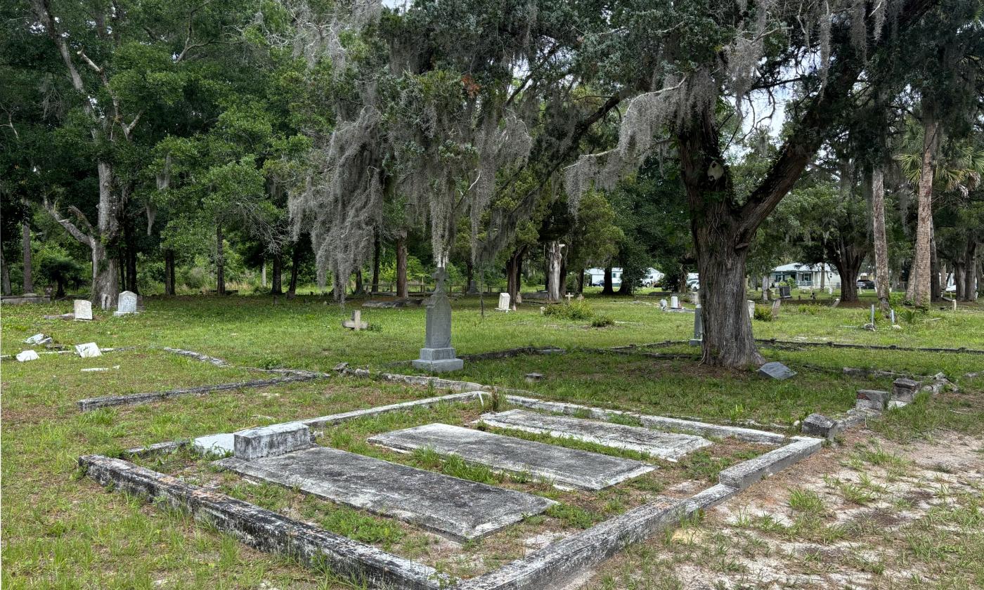 View of the Pinehurst and San Sebastian Cemeteries. A variety of grave markers spread beneath palm, cedar, and oak trees.