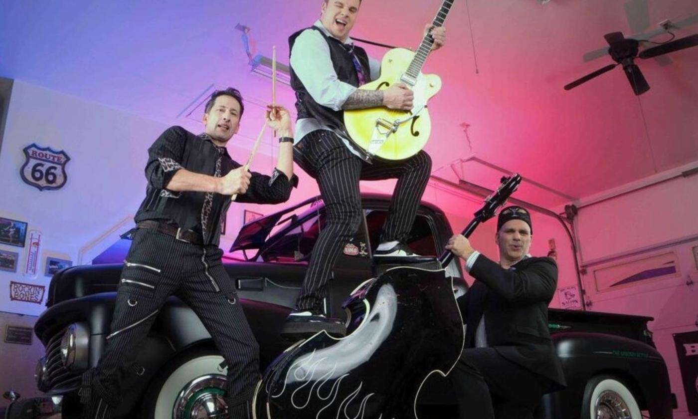 Bandmates from Rock This Town - Rockabilly Riot pose with their guitars near a classic truck.