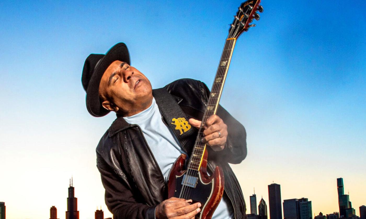 Ronnie Baker Brooks wears a black leather jacket and matching hat while playing the guitar. 