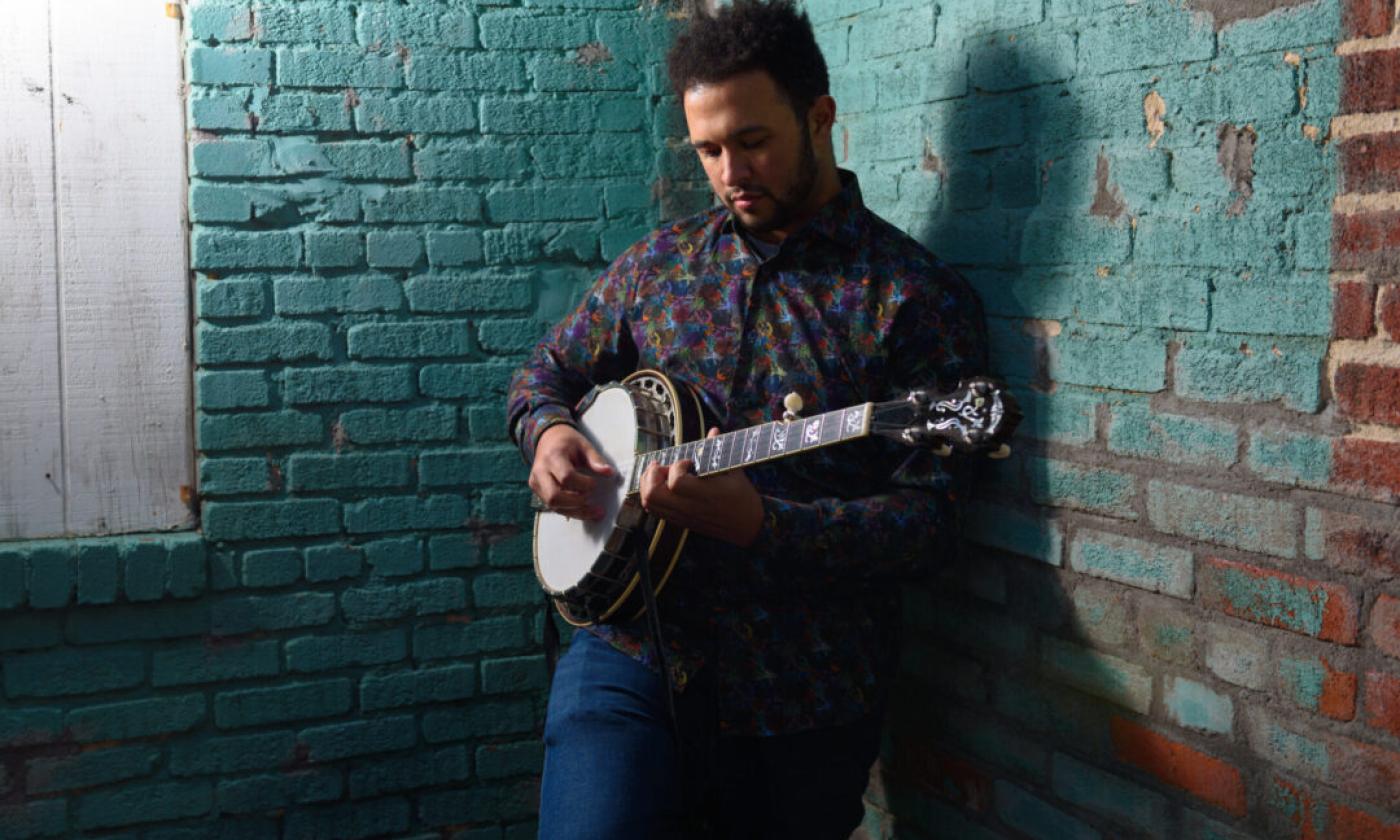 Tray Wellington strums his banjo in front of a brick wall.