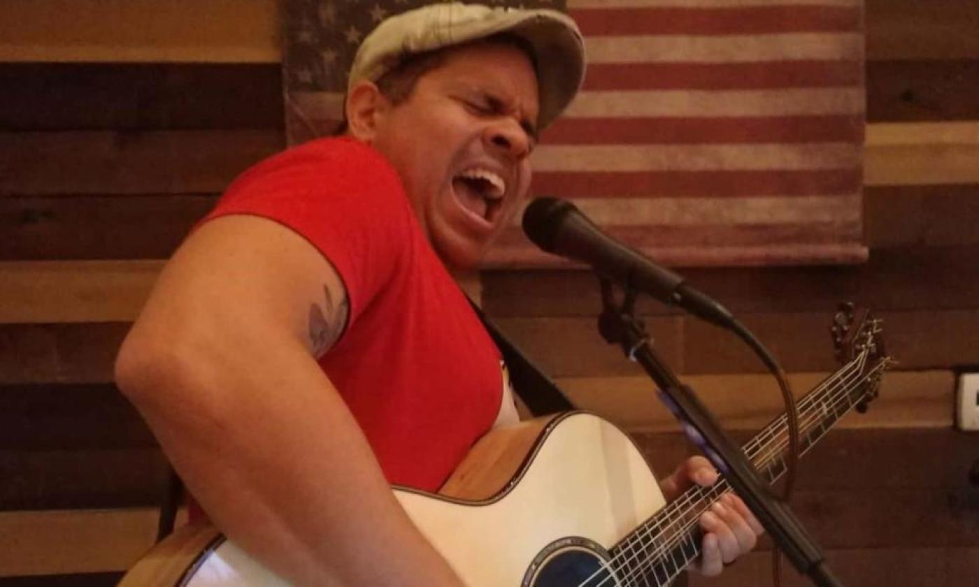 Guitarist Edwin Barbosa singing onstage with guitar in red shirt with cap.