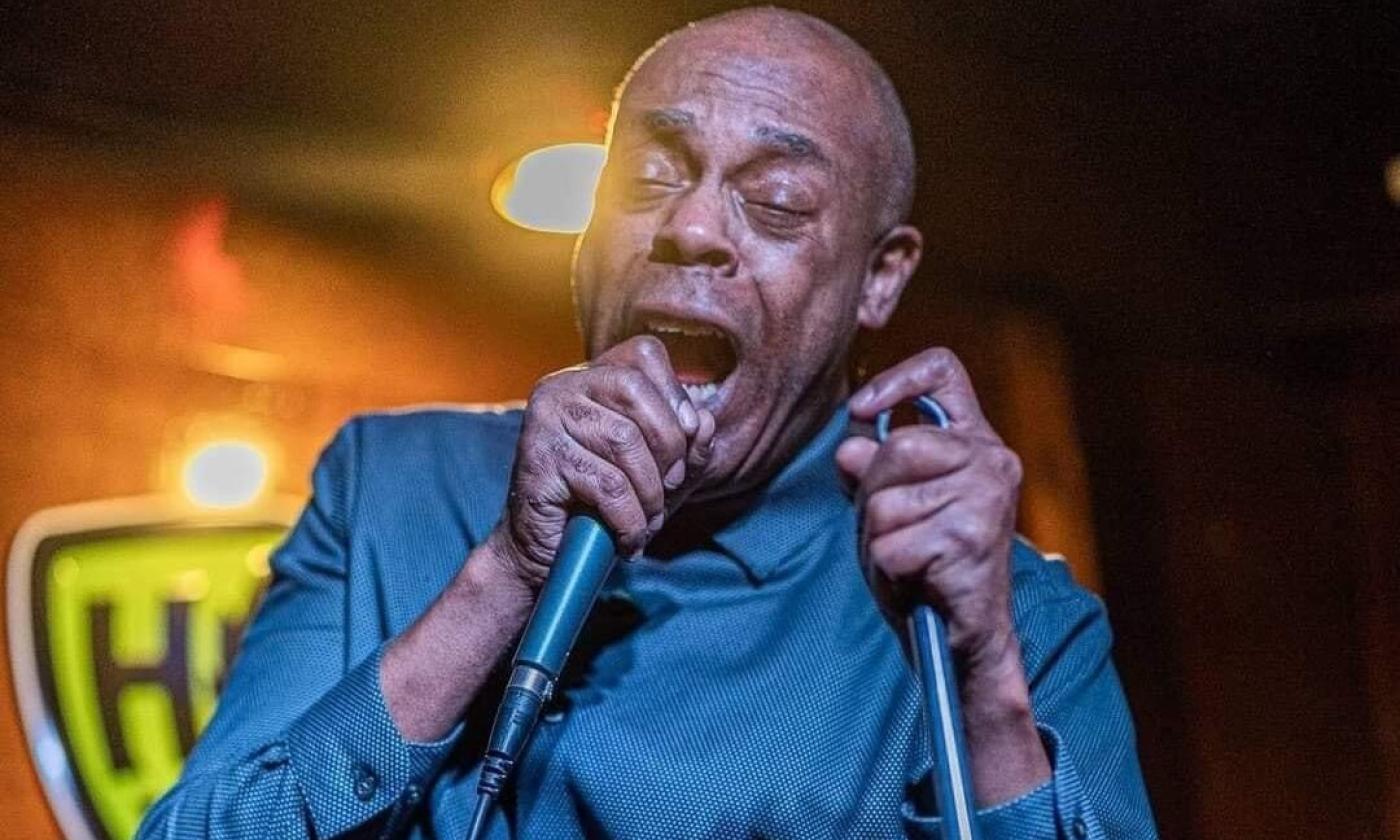 Michael Winslow wears a blue shirt while speaking with a microphone pressed to to his lips.