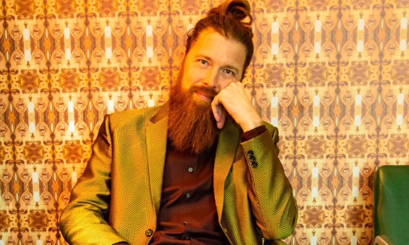 Shane Mauss sits in a chair and poses with a brown blazer and top in front of wallpaper with strange faces in the form of a pattern.