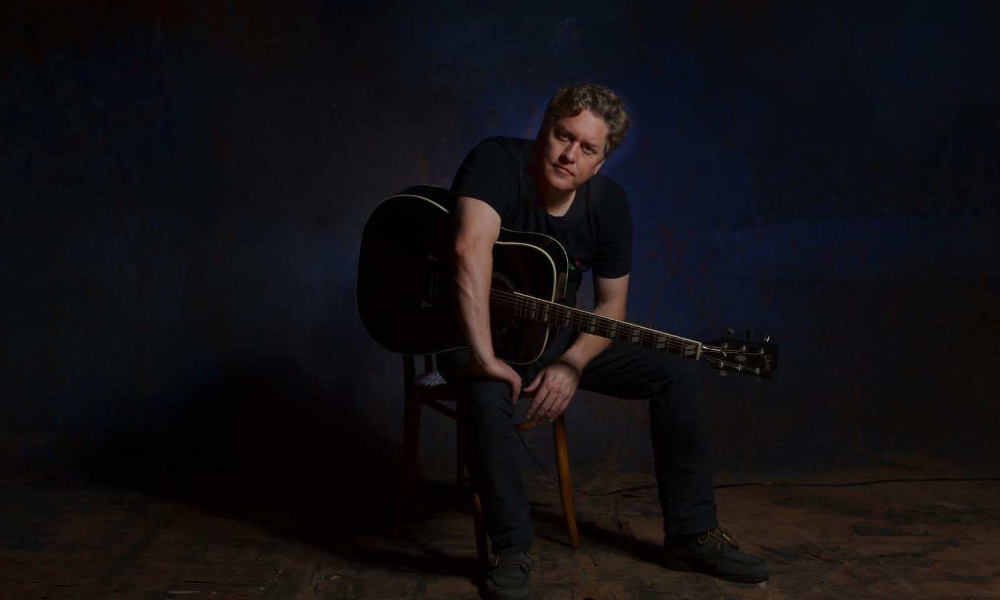 Shawn Camp, singer and guitarist, sitting with his guitar on a dark stage