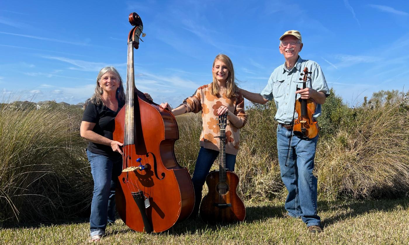 Members of a bluegrass and folk trio, standing in a field with their instruments under a blue sky