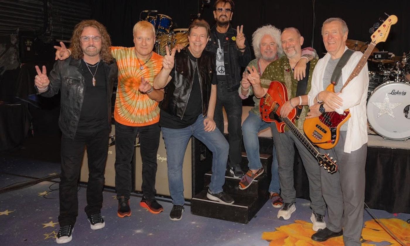 Bandmates from Ringo Starr and His All-Starr Band pose on stage with their musical instruments.