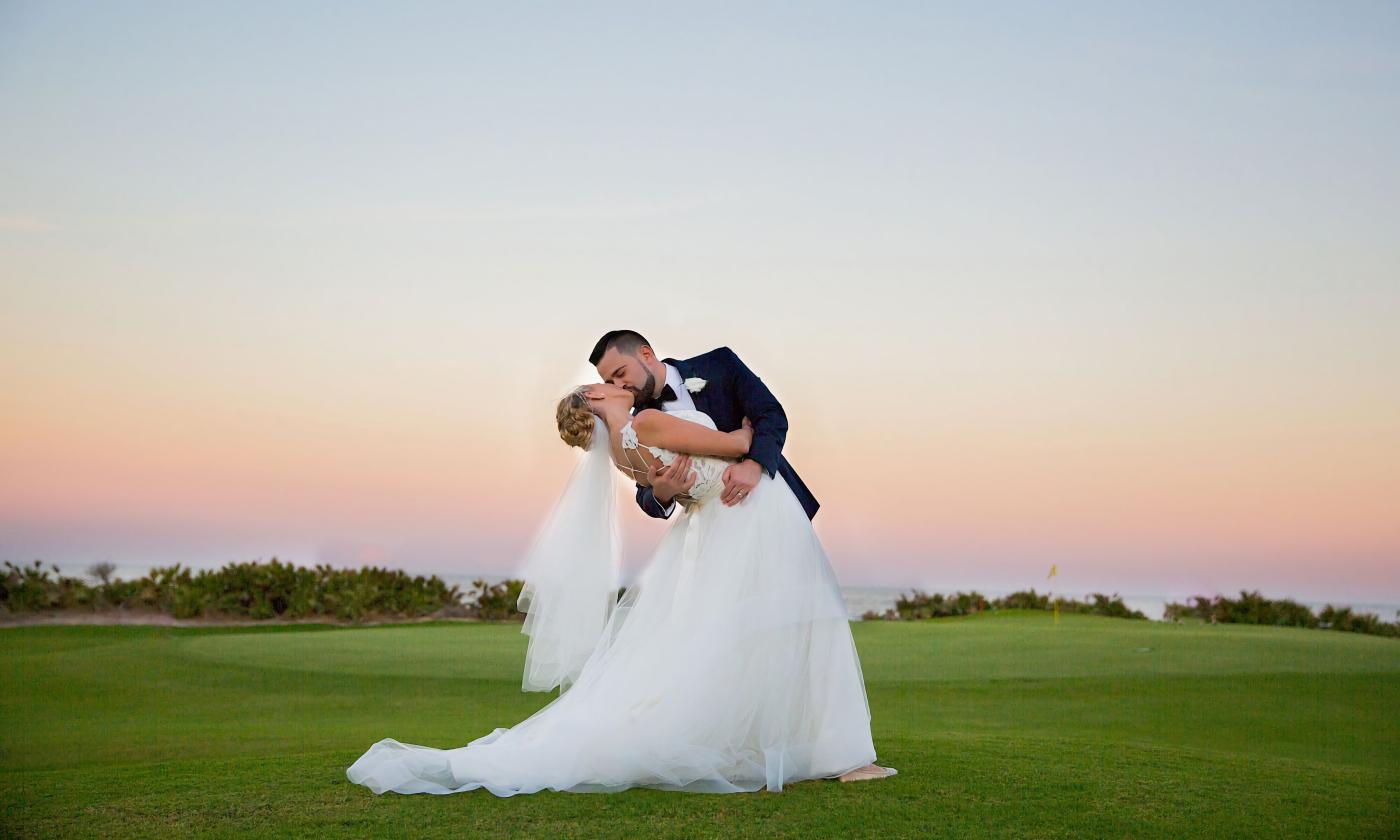 A bride and groom kiss near a green on a golf course at sunset