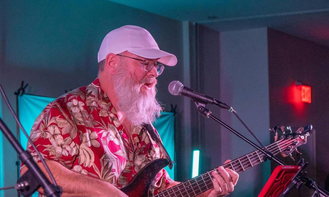 Marshall Bailey in white cap and Hawaiian shirt, singing at a microphone with his guitar