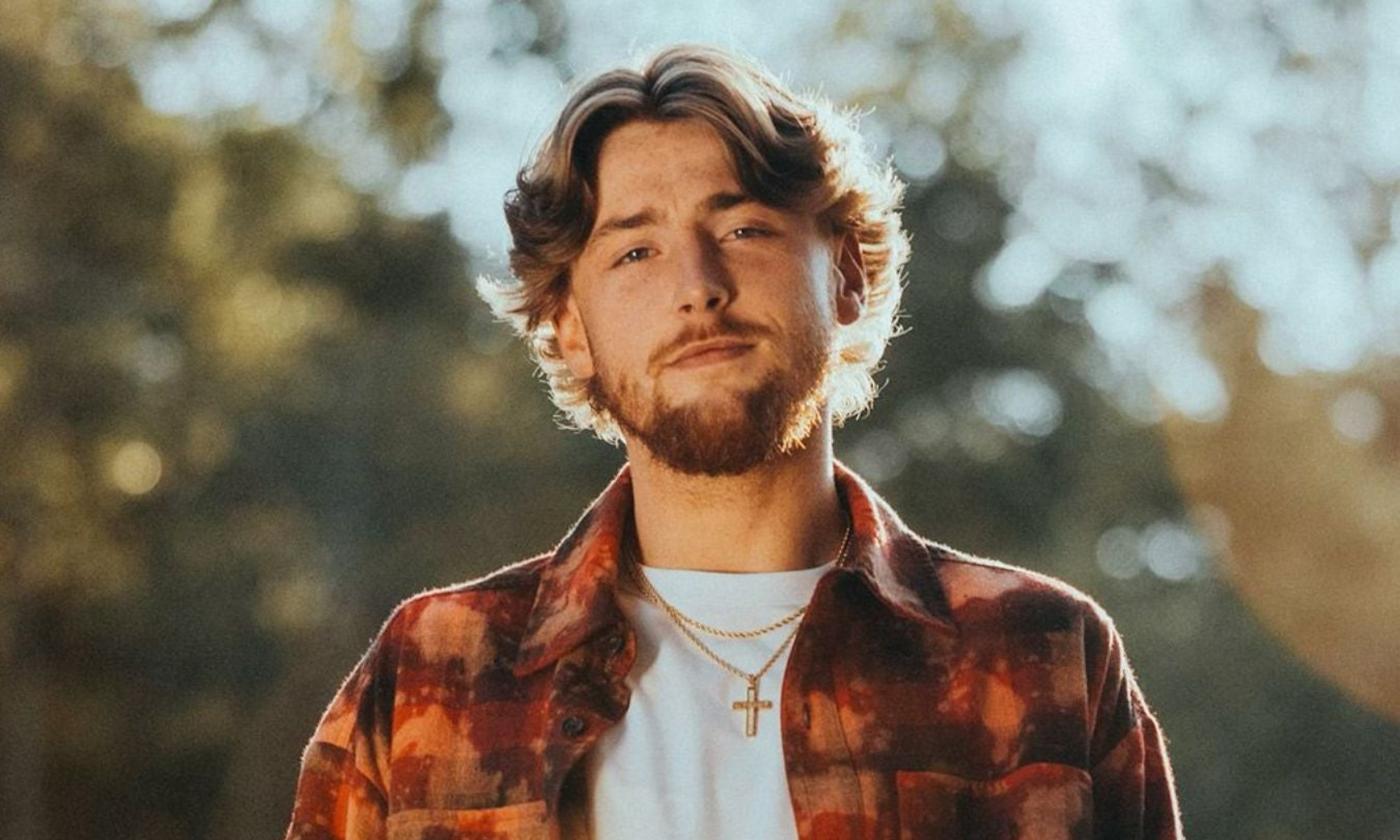 Bailey Zimmerman wears a plaid shirt and smiles in front of a nature-themed backdrop.