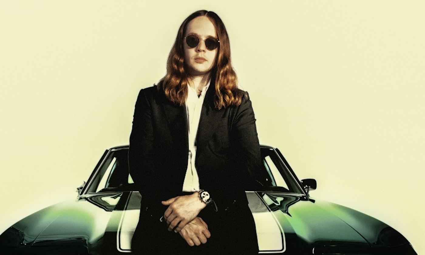 Billy Strings wears black glasses while posing in front of a dark green vehicle.