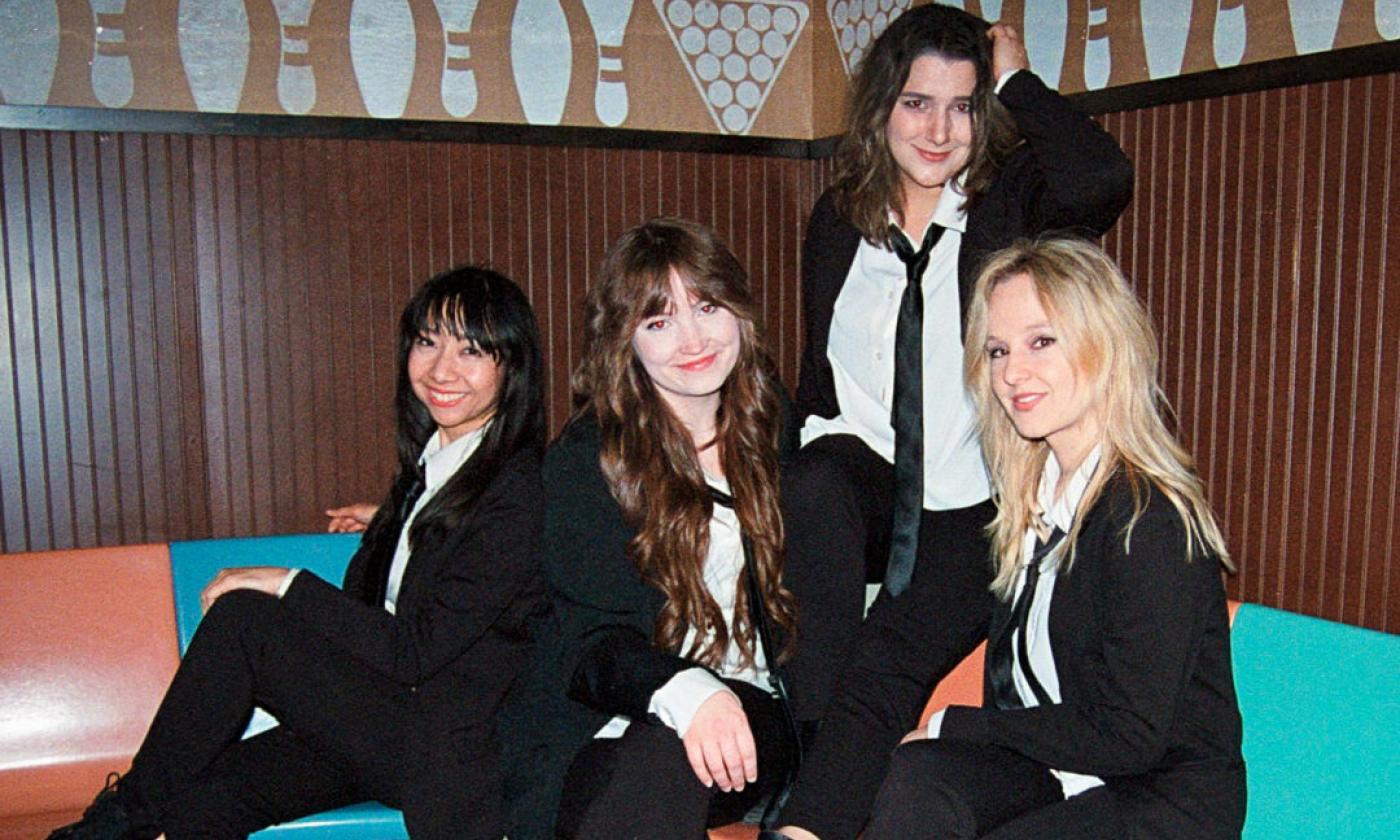 Bandmates from Penny Lane wear black pants and matching blazers while posing in front of a brown and black backdrop with textures and patterns. 