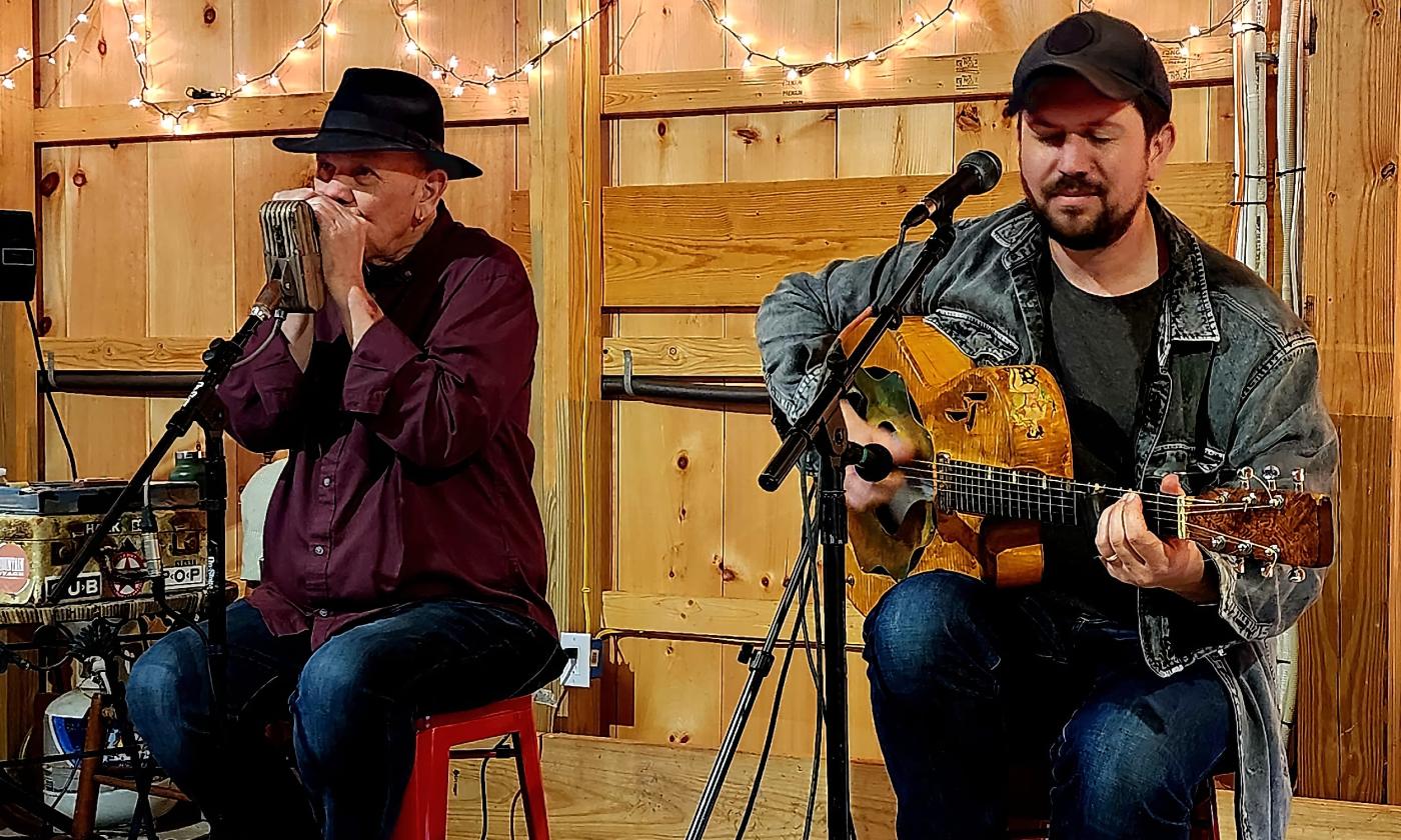 Two musicians on stage in a barn, both seated, the one on the left is playing the harmonica; the one on the right, the guitar