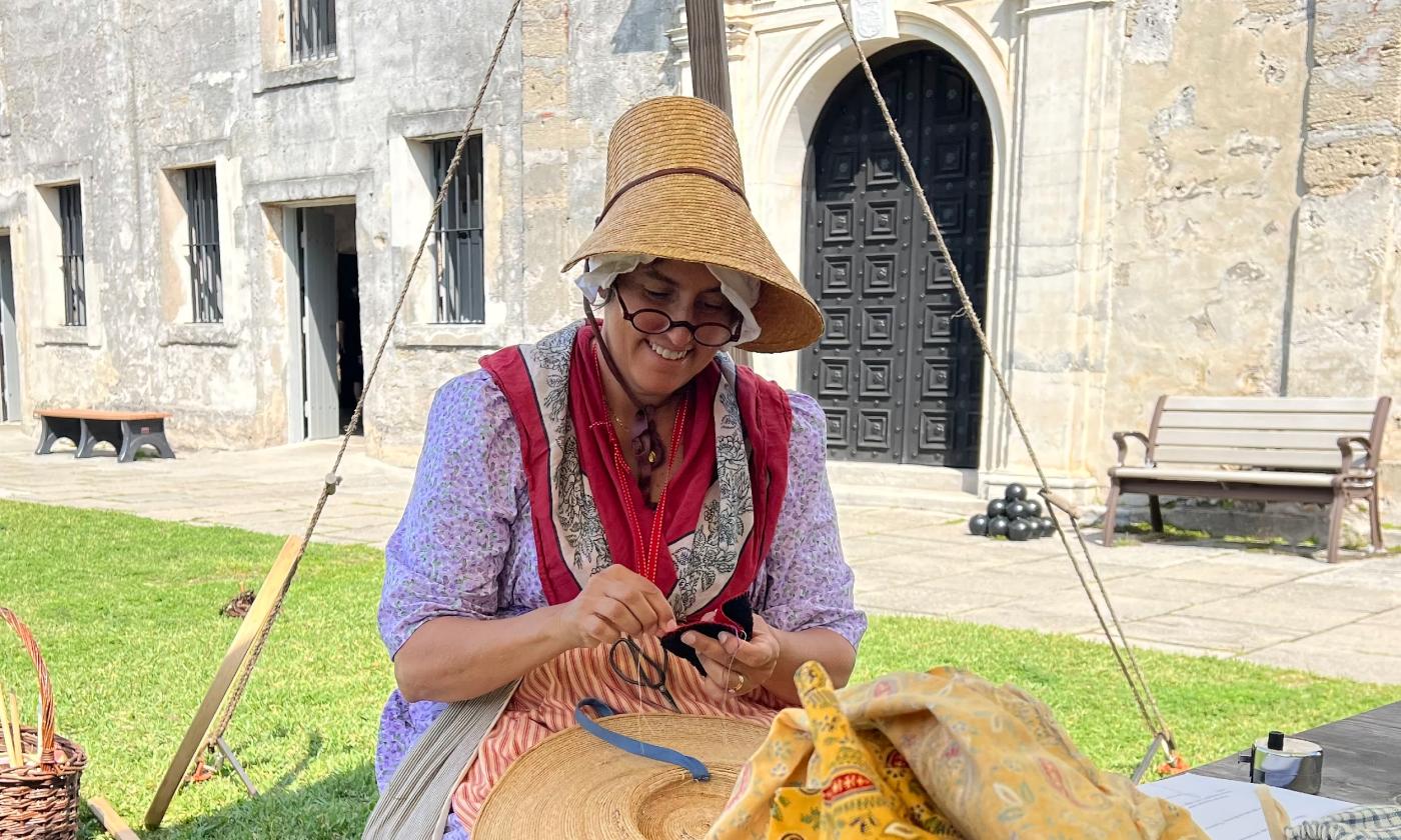 A woman dressed in colonial clothing and a straw hat had sews clothing at the Castillo de San Marco