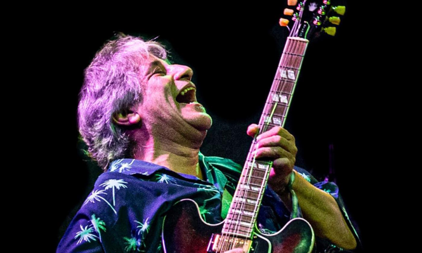 Chris Cain smiles and sings with his guitar in front of a black backdrop. 