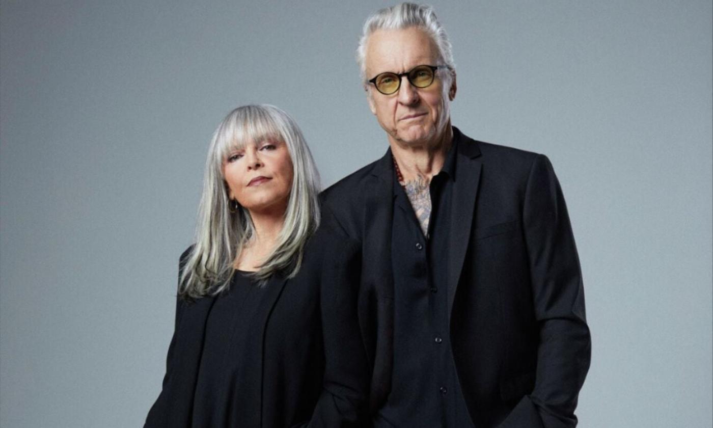 Pat Benatar & Neal Giraldo wear black and pose in front of a gray backdrop.