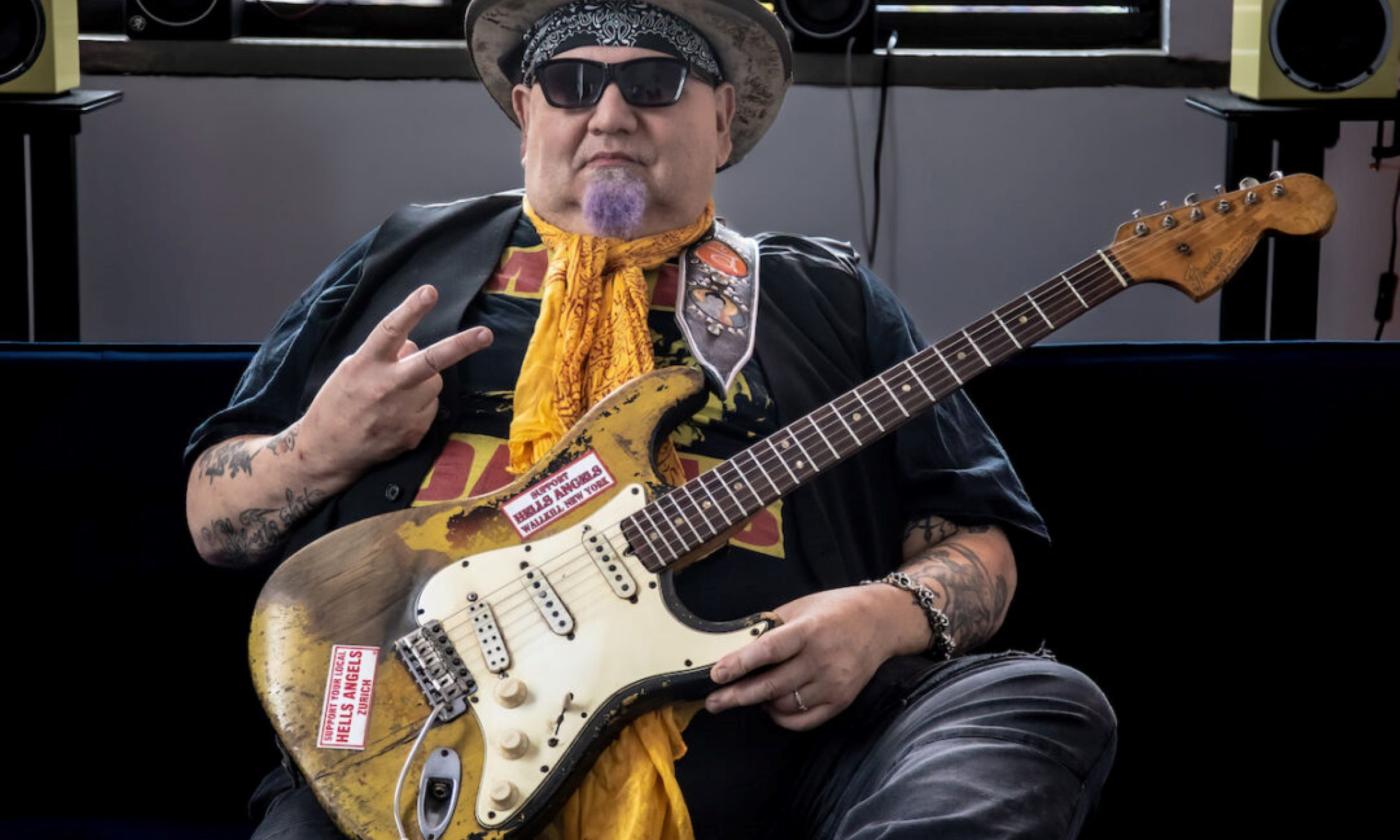 Popa Chubby sits on a chair and poses with his guitar.