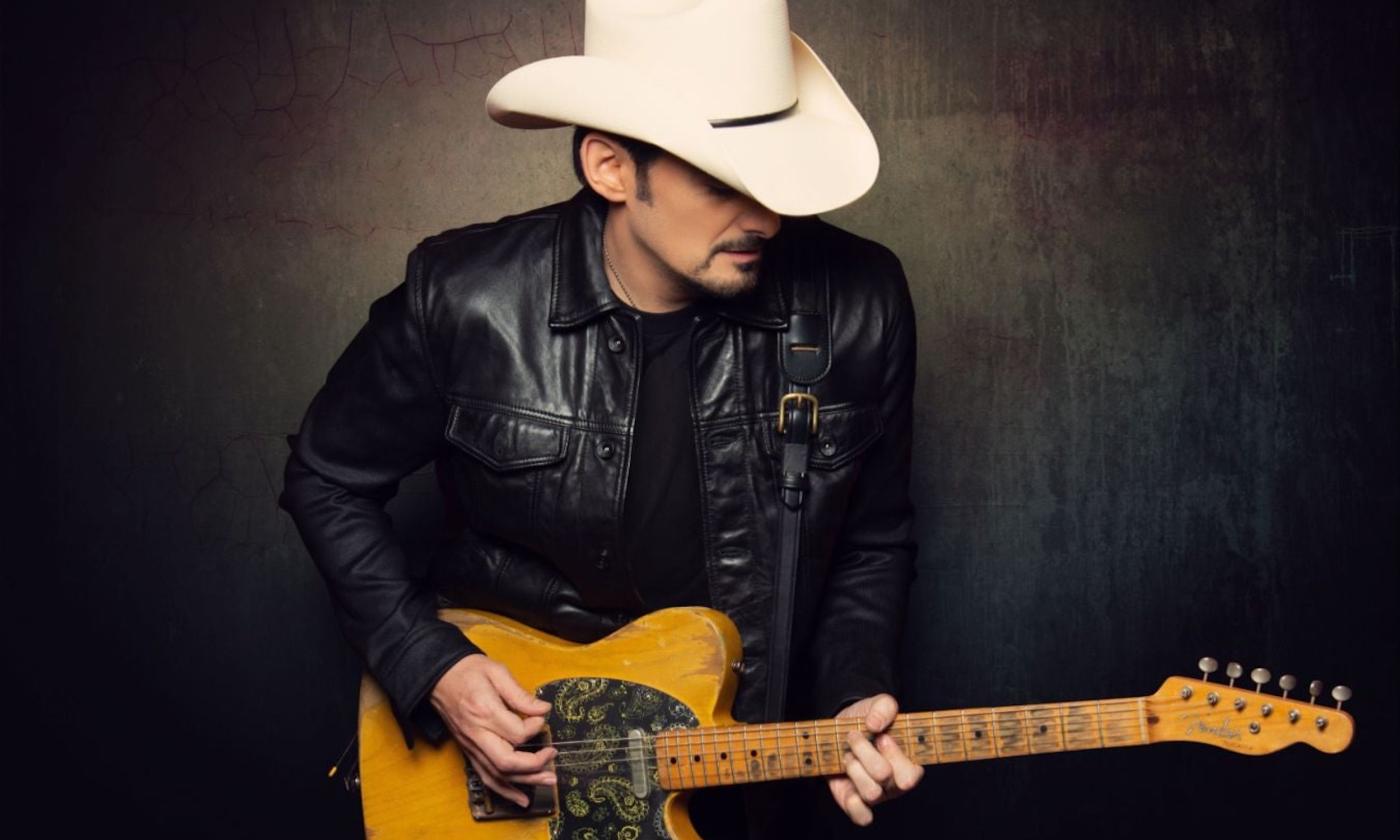 Brad Paisley wears a white cowboy hat and leather jacket while clutching his guitar in front of a black backdrop. 