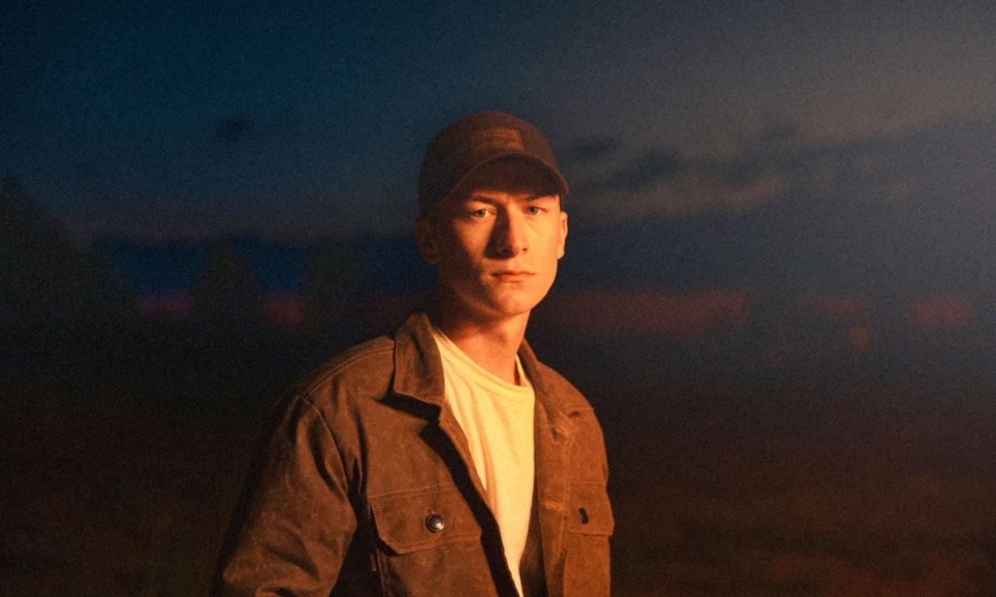 Sam Barber wears a black cap and brown jacket while posing in front of a black and blue backdrop. 