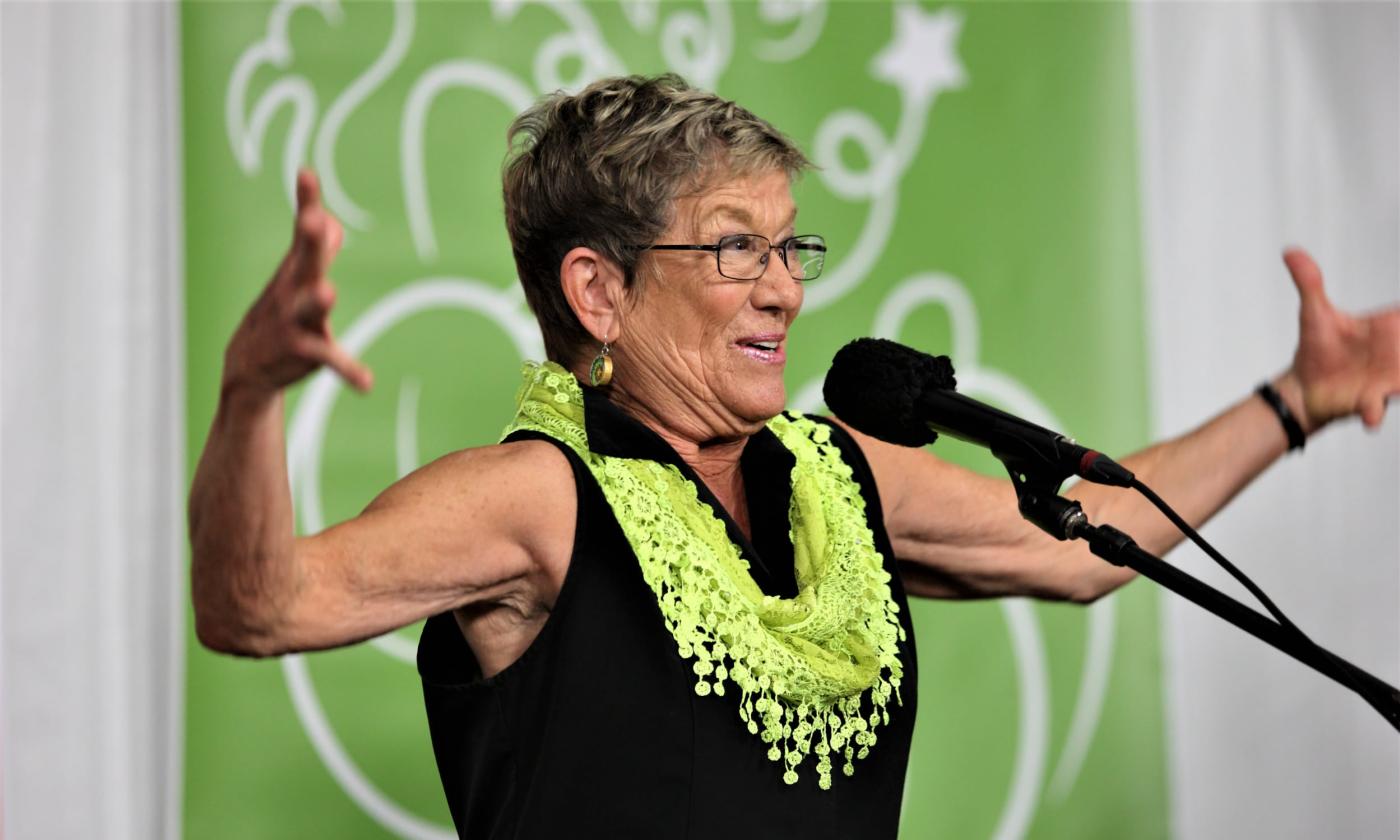 Story teller Pat Huge on stage in black with a green scarf, with her arms spread wide