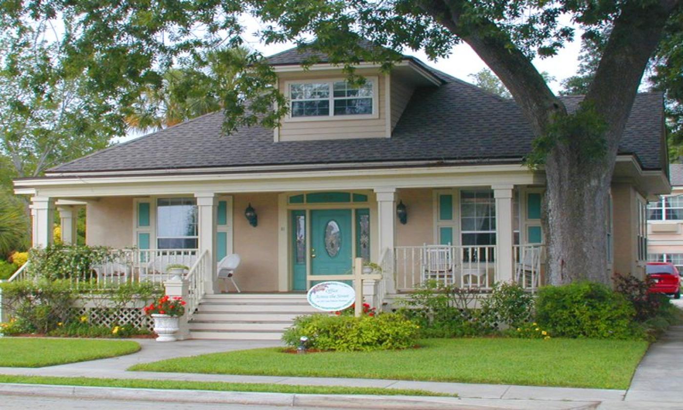 A pink two-story home owned by Cozy Inn St Augustine. Its wrap-around porch holds white wicker rocking chairs. Flower bushes and oak trees surround it