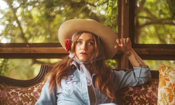 Sierra Ferrell, wearing a straw hat, and sitting on a flowered sofa, in front of a wall of windows