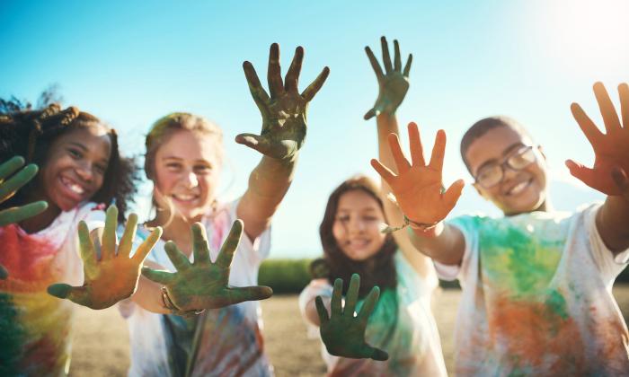 A group of grinning children hold their hands to the camera to show paint stained, which are also on their clothes