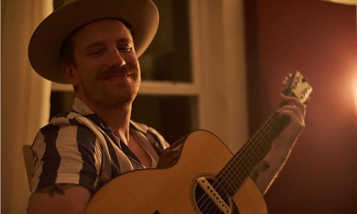 Singer and songwriter Chris Rudasill, wearing a striped shirt and a hat, playing guitar in a softly lit room