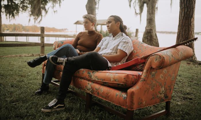 The duo that is the band Coyboi, sitting on a sofa, under palm trees, near the river