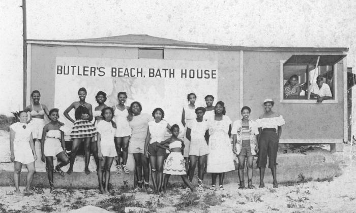 B&W a group of young Black women pose in front of a building, mural and sign that reads "Butler's Beach, Bath House." An older couple leans out the window to smile for the camera.
