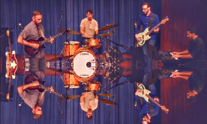 Four band members perform in front of a blue curtain. Their images are reflected on the bottom half of the image.