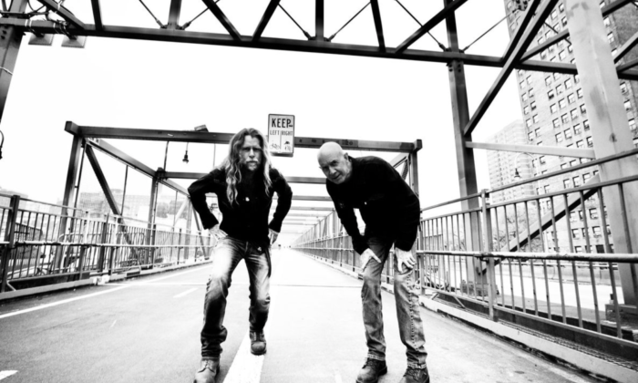 A black and white photograph of Grayson Capps and Corky Hughes posing on a bridge with beams and a walkway as a backdrop. 