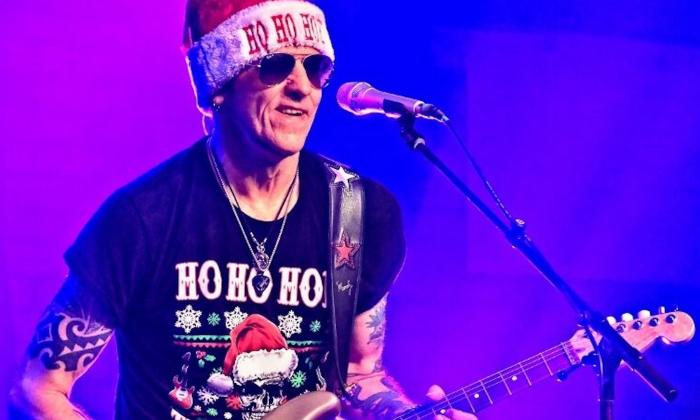 Gary Hoey strums his guitar on stage while wearing a bright red Christmas hat. 