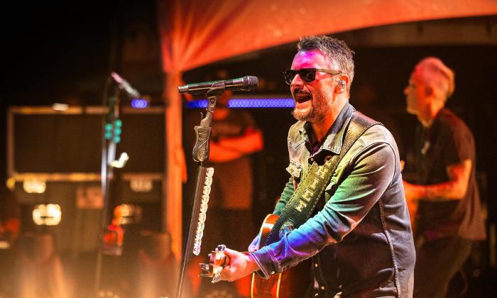 Eric Church on stage with his band, singing and playing guitar under a red light