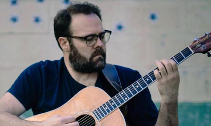 Josh Daniel strums his guitar in front of a gray and teal backdrop. 