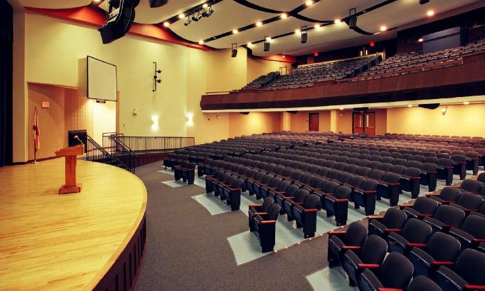 An auditorium, with two tiers, a light wood stage, and a scree for titles