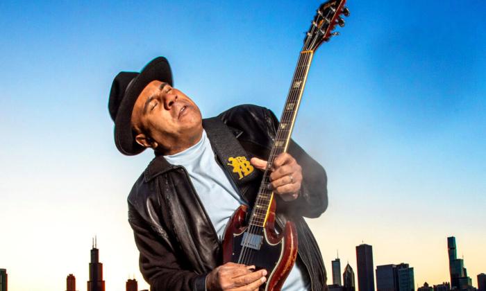 Ronnie Baker Brooks wears a black leather jacket and matching hat while playing the guitar. 